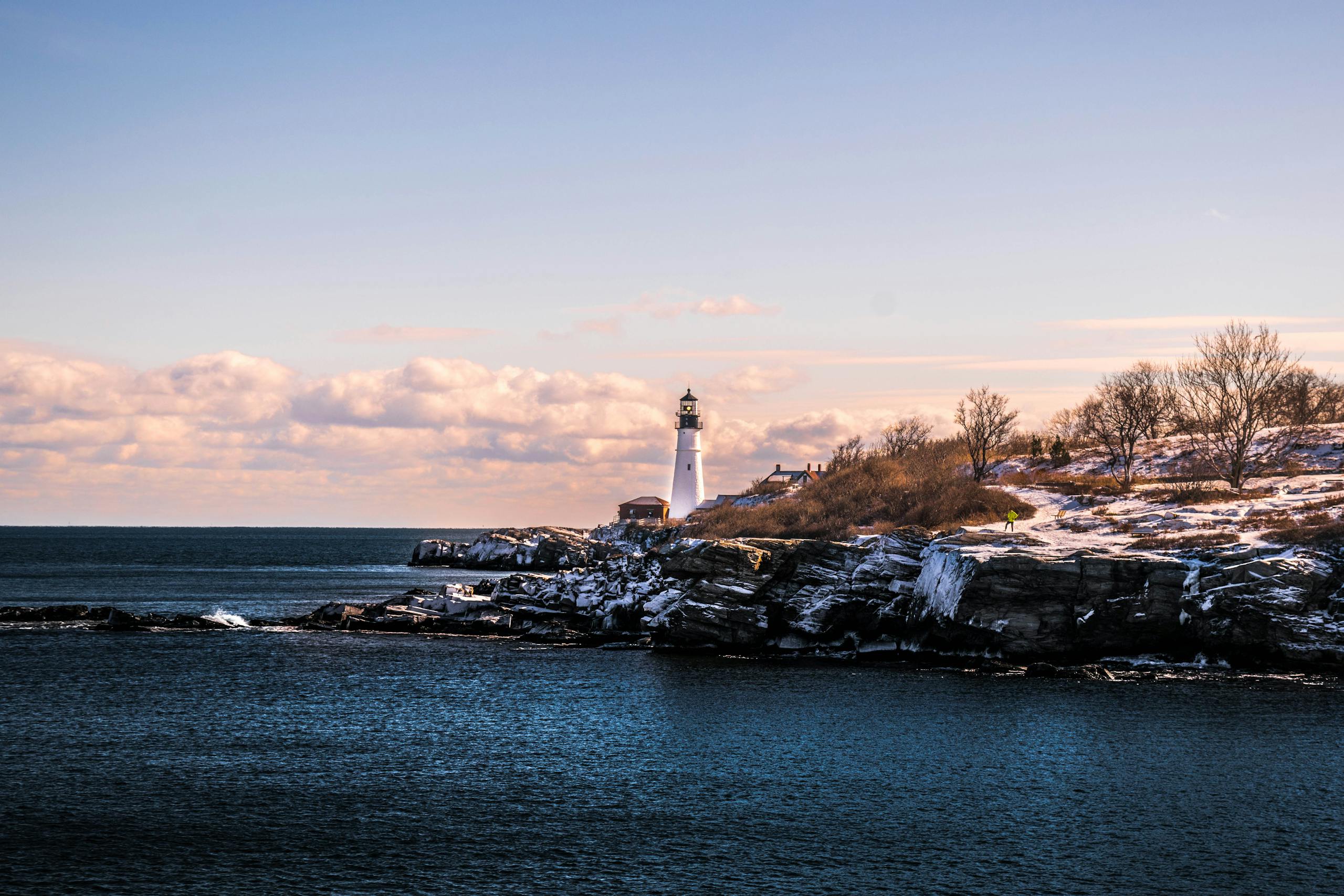 A picturesque winter view of a lighthouse on the rocky Maine coastline under a bright sky.