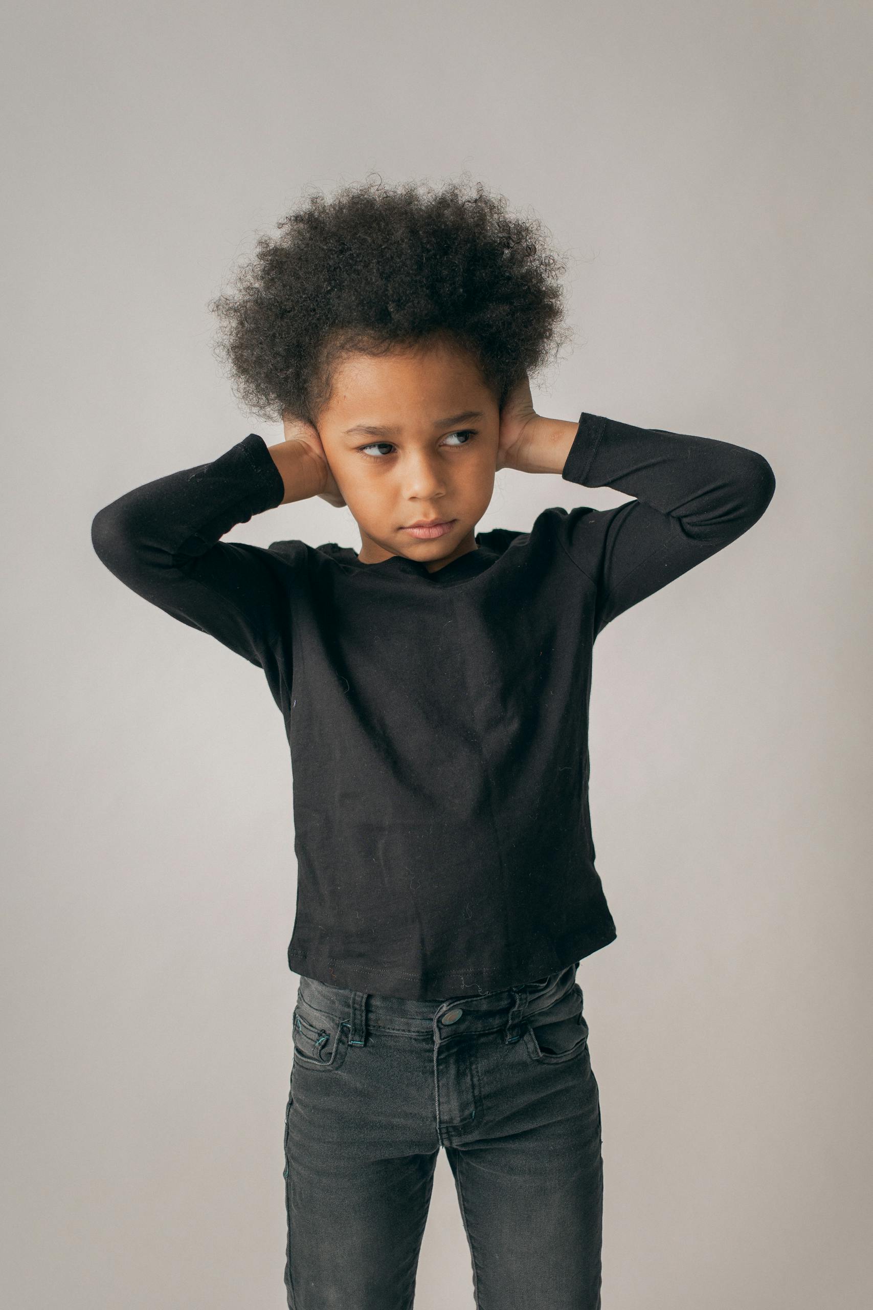 Desperate African American girl in black outfit and Afro hairstyle looking away while covering ears on white background in room