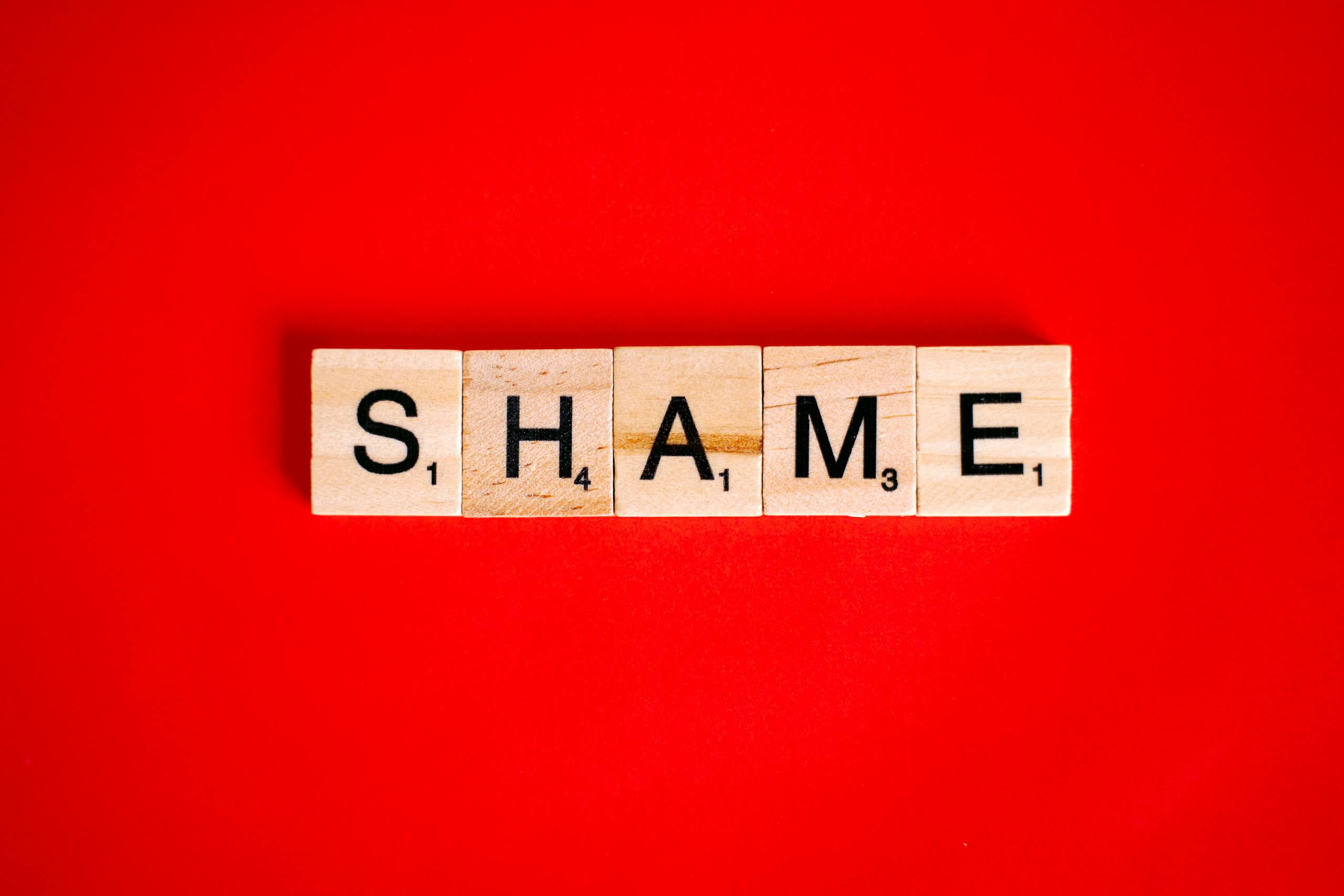 Scrabble tiles spelling the word 'Shame' on a vibrant red backdrop.
